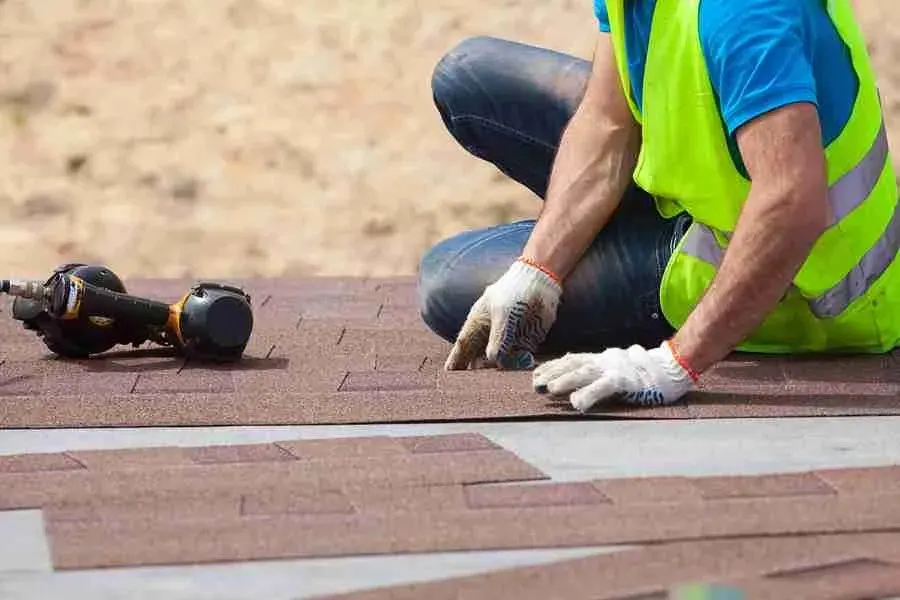 Roofer in a safety vest kneeling on a shingle roof with a nail gun