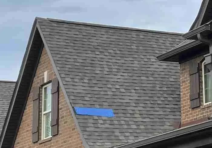 Brick house with a gray shingle roof and a blue tarp strip marking a repair
