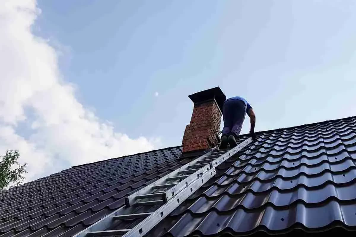 Roofer climbing a ladder on a dark metal tile roof toward a brick chimney