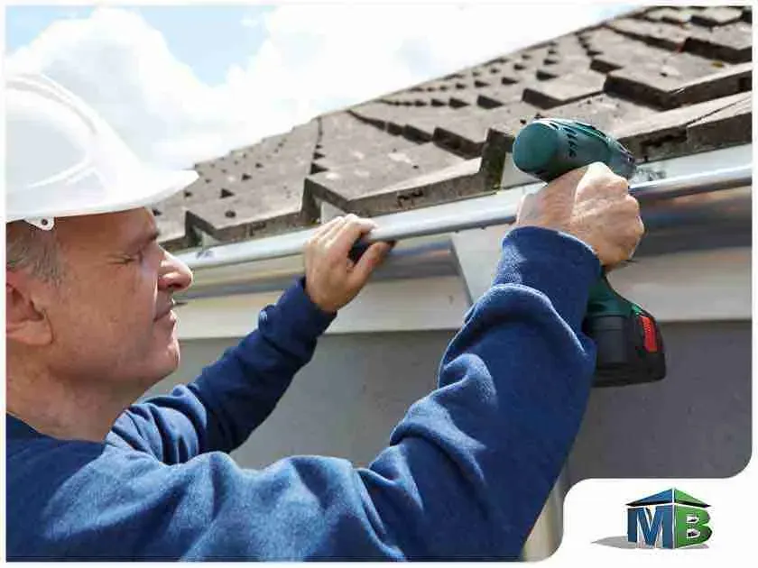 Worker in a hard hat drilling a gutter bracket along a shingle roof edge