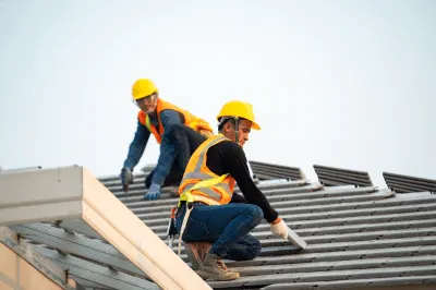 Two roofers in hard hats and safety vests working on a metal roof