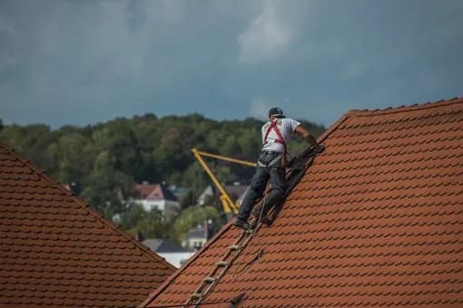 Roofer in a harness climbing a ladder on a red tile roof