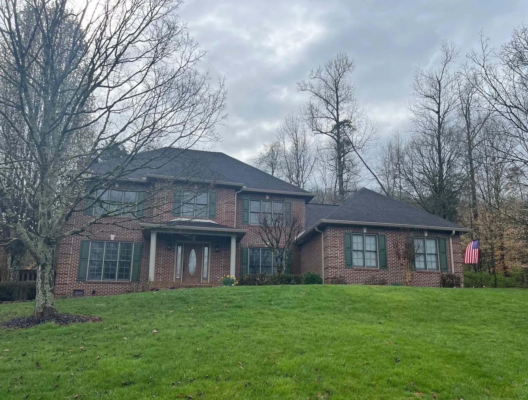 A large brick two-story house with a dark shingle roof and green shutters, surrounded by a green lawn and bare trees under an overcast sky