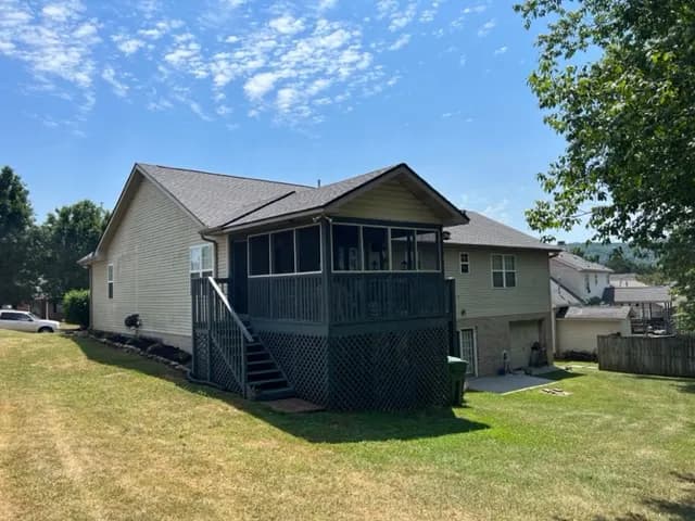 A single-story house with a gray shingle roof, screened-in porch deck, and a landscaped yard on a sunny day