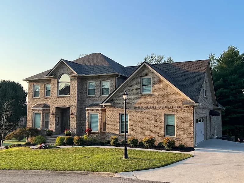 A large two-story brick home with a dark gray shingle roof, landscaped front yard, and paved driveway under a clear blue sky