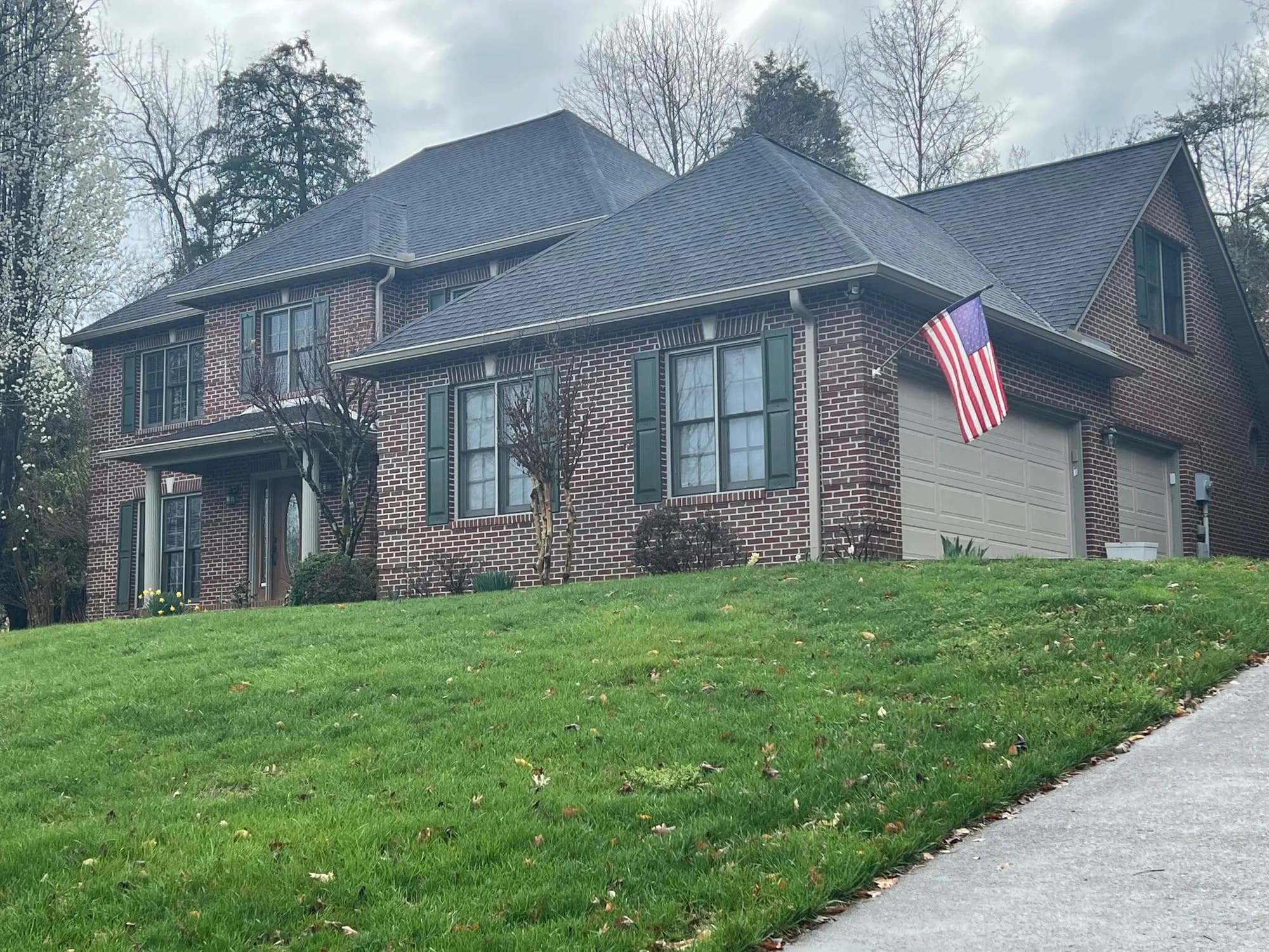 A two-story brick house with a gray shingle roof, green shutters, and an American flag displayed near the garage, viewed from the front lawn