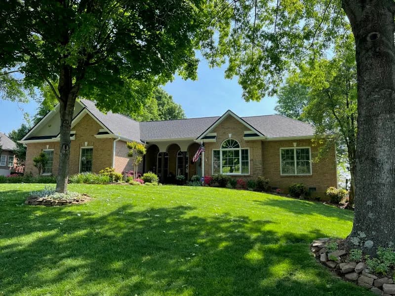 A single-story brick home with a well-manicured lawn, mature trees, and a shingled roof