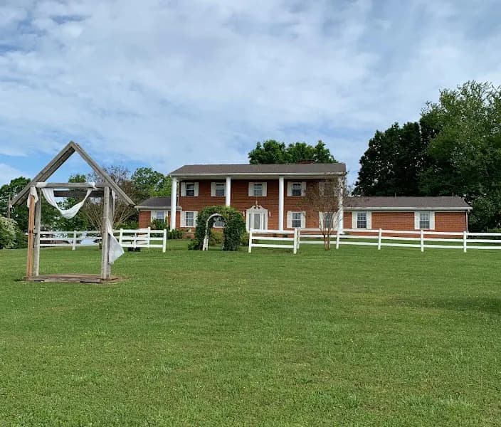 A large two-story brick colonial-style house with white columned porch entrance, surrounded by a green lawn and white split-rail fencing, viewed from across a g