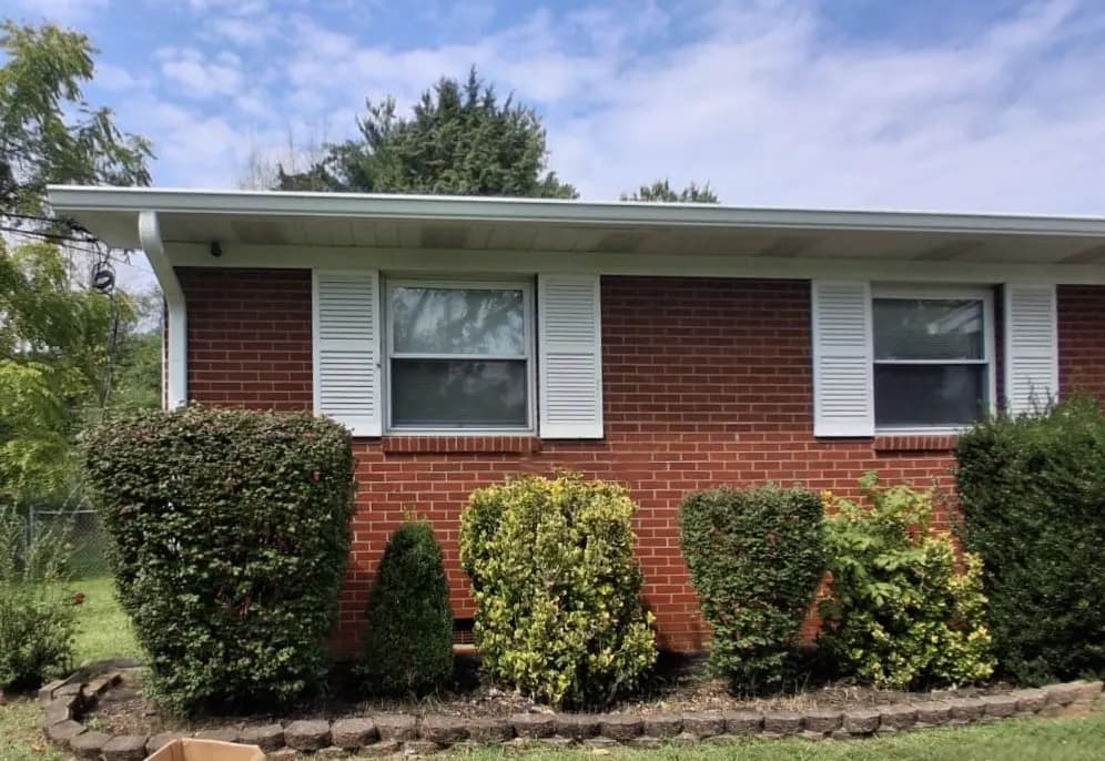 Exterior view of a brick ranch-style home with white shutters, gutters, and landscaped shrubs, showing the roofline and eaves