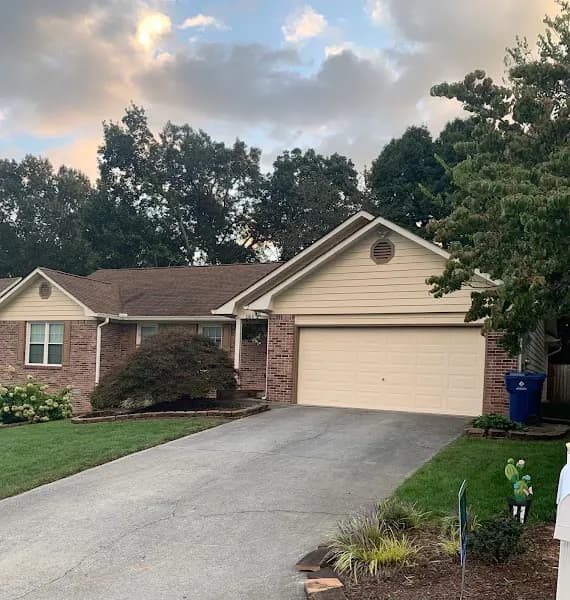 A single-story brick and beige-sided ranch home with a two-car garage and roof, photographed from the driveway at dusk