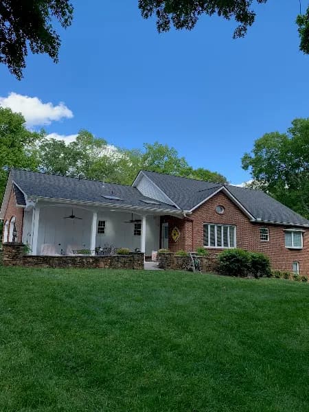 A brick ranch-style house with a shingled roof and covered front porch, surrounded by a green lawn and trees under a blue sky