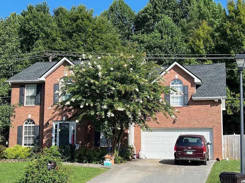 A two-story brick house with a gray asphalt shingle roof, white garage door, and a maroon minivan parked in the driveway