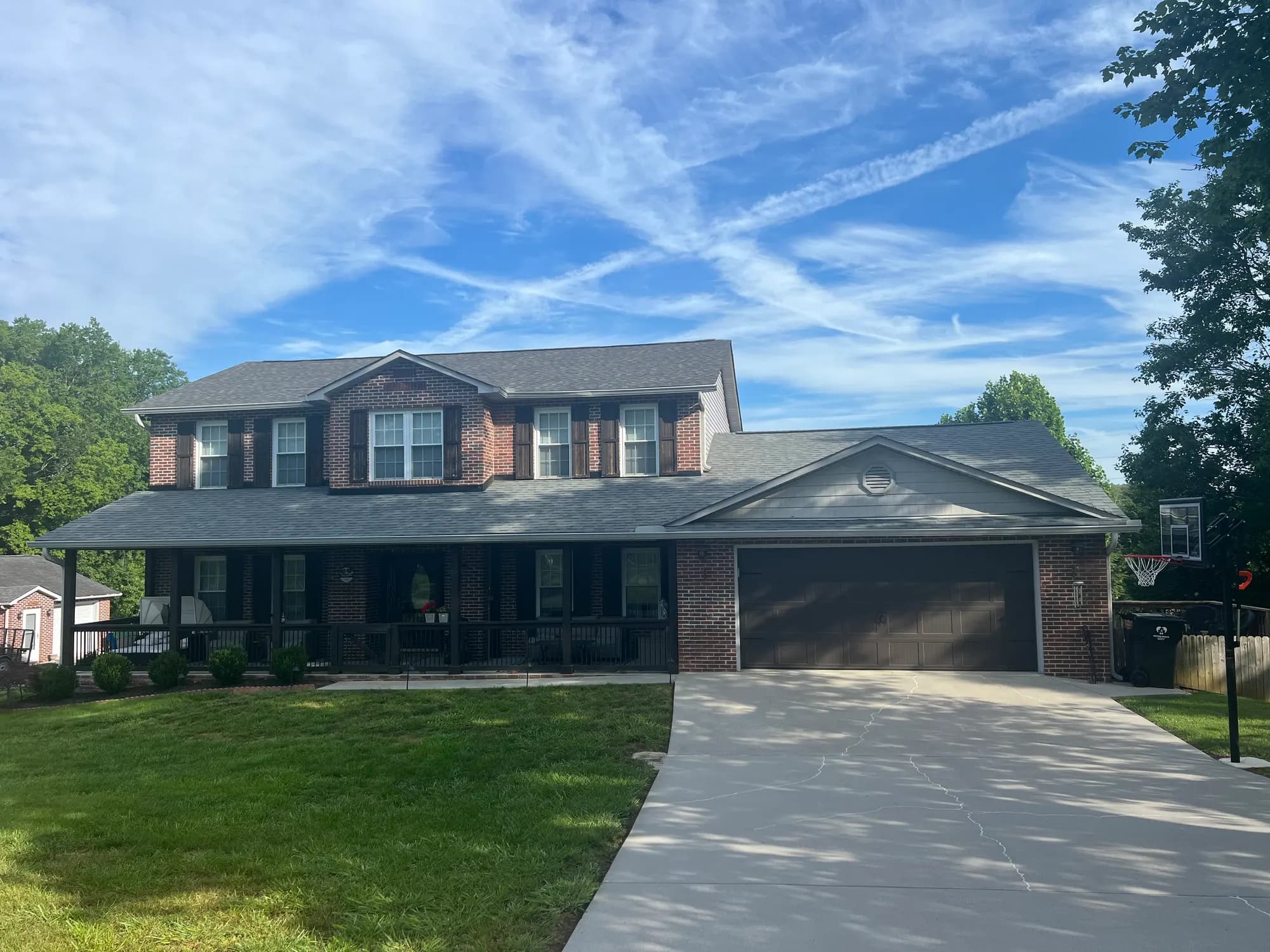 A large two-story brick house with a new gray shingle roof, wraparound porch, and attached two-car garage under a bright blue sky