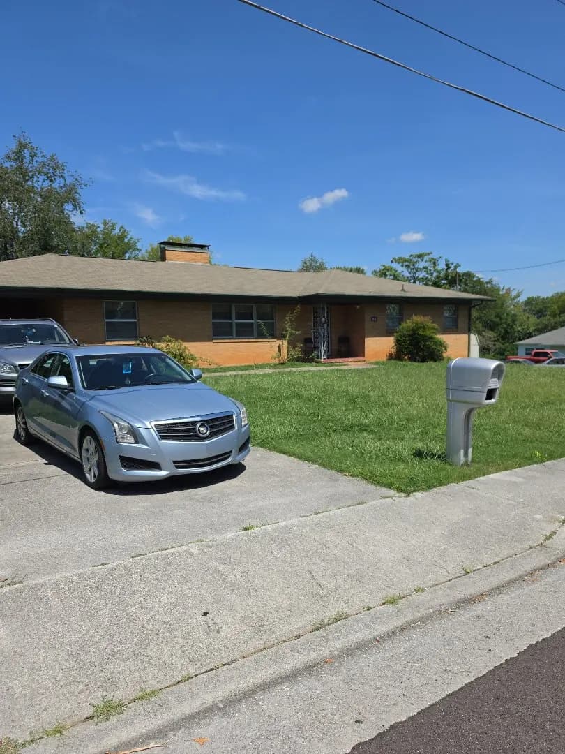 A single-story brick ranch-style home with a shingled roof, parked cars in the driveway, and a mailbox on the front lawn