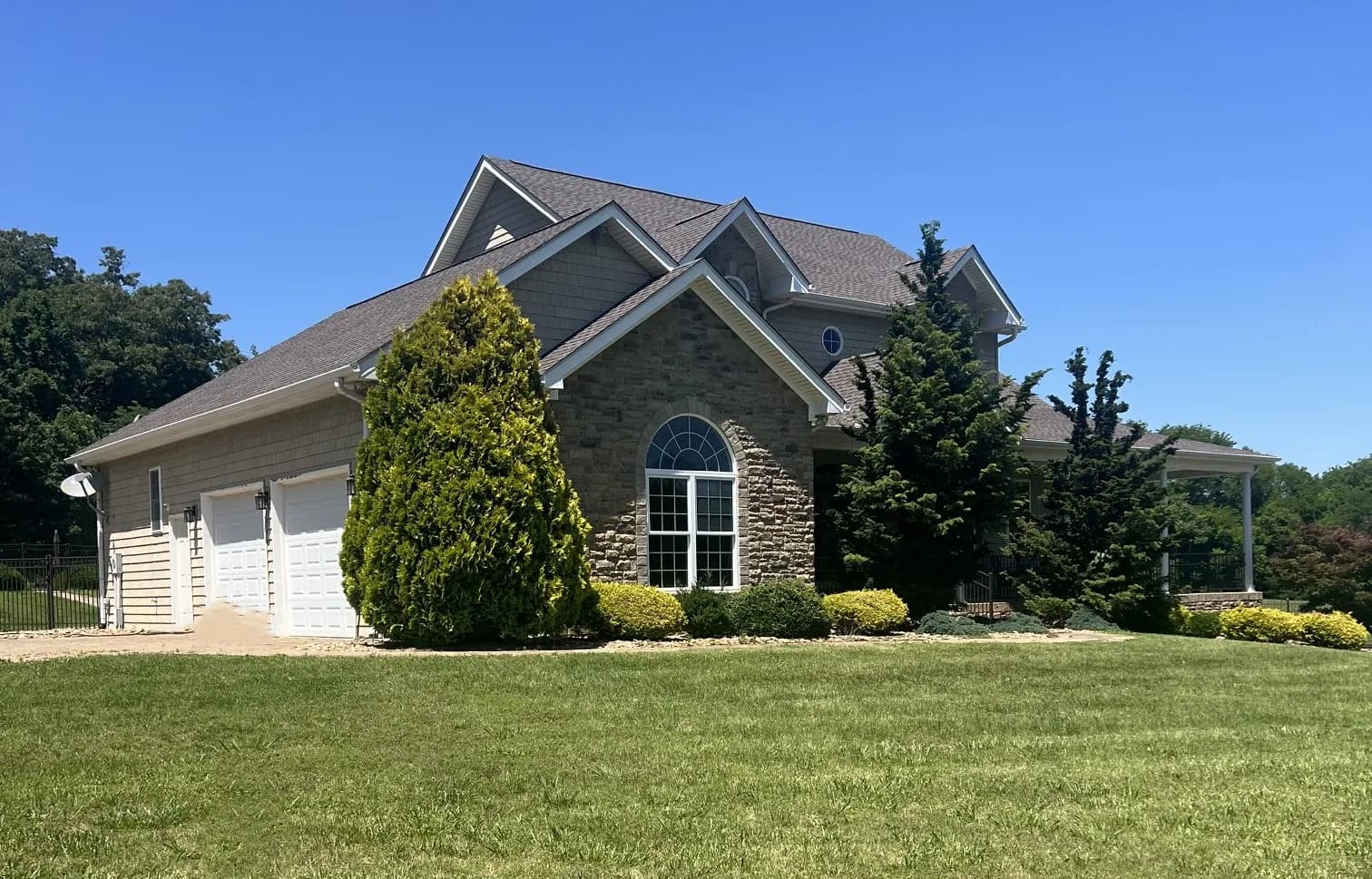 A stone and shingle-sided two-story house with a landscaped front yard, mature evergreen trees, and an asphalt shingle roof under clear blue sky