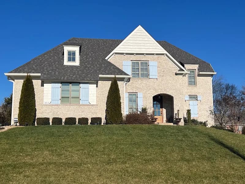 A two-story stone and brick home with a dark gray shingle roof, blue shutters, and a well-maintained lawn under a clear blue sky
