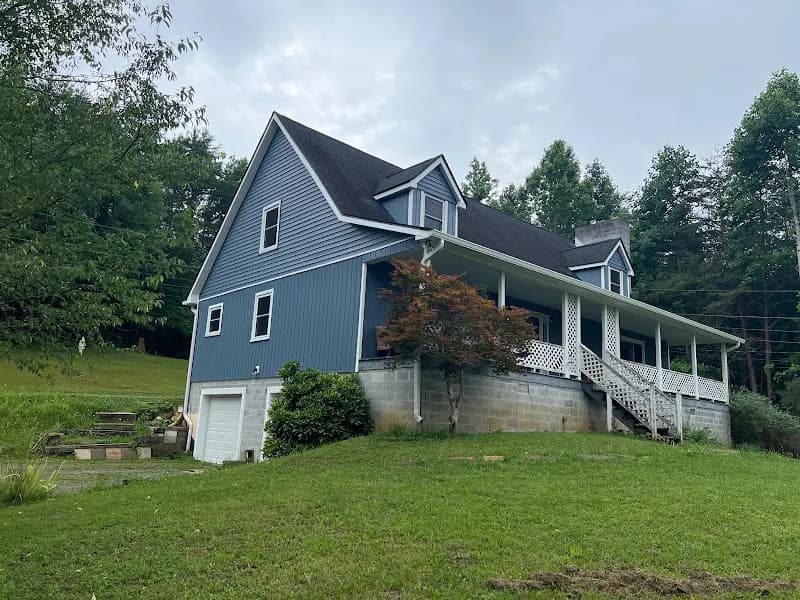 A two-story blue house with a gray shingle roof, dormer windows, wraparound porch, and stone foundation, surrounded by a grassy yard and trees