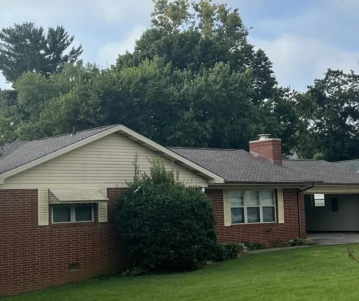 Exterior view of a brick ranch-style home with a beige-shingled roof, chimney, and carport, surrounded by trees and lawn