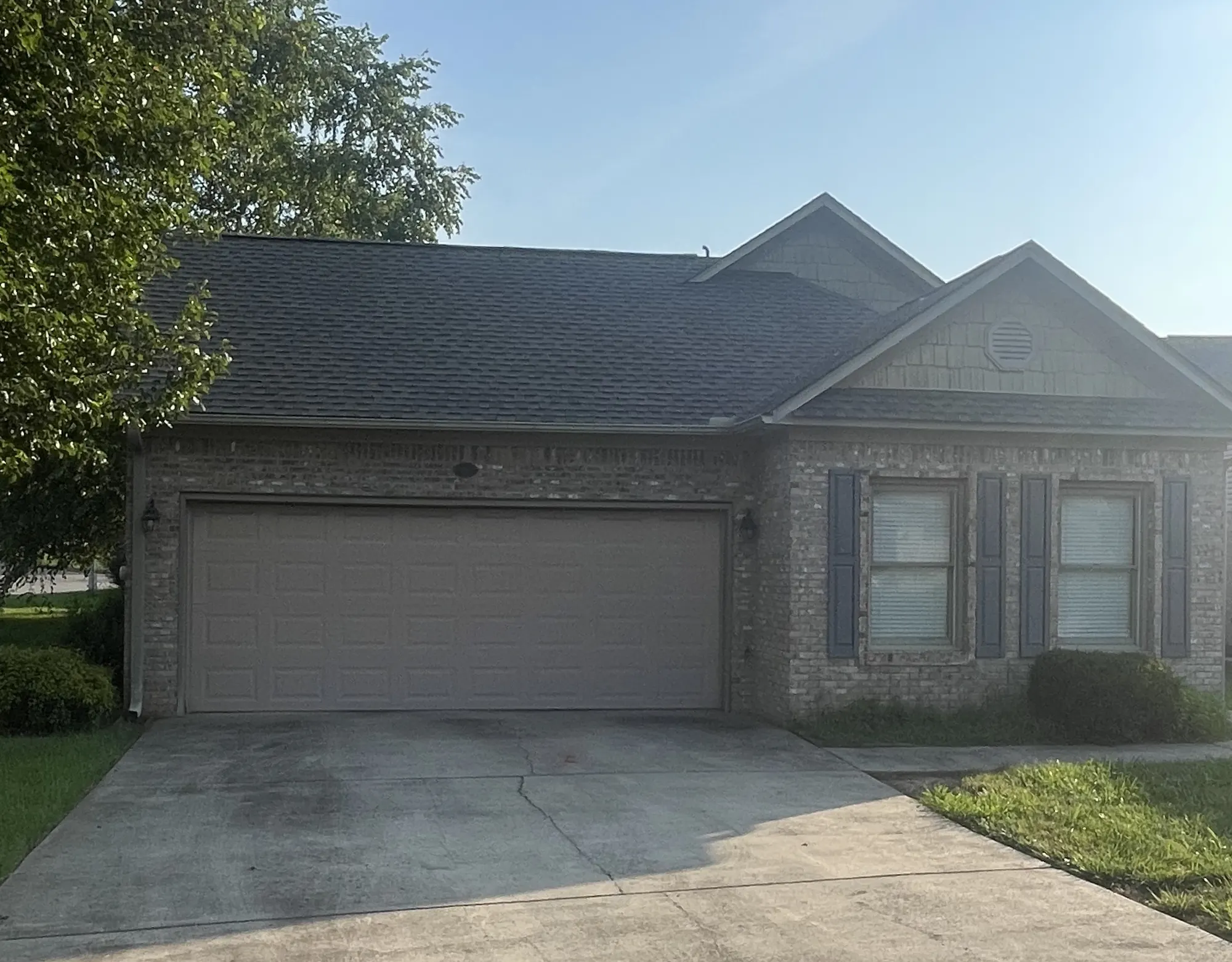 Front exterior view of a single-story brick home with a shingled roof, two-car garage, and a large tree in the front yard