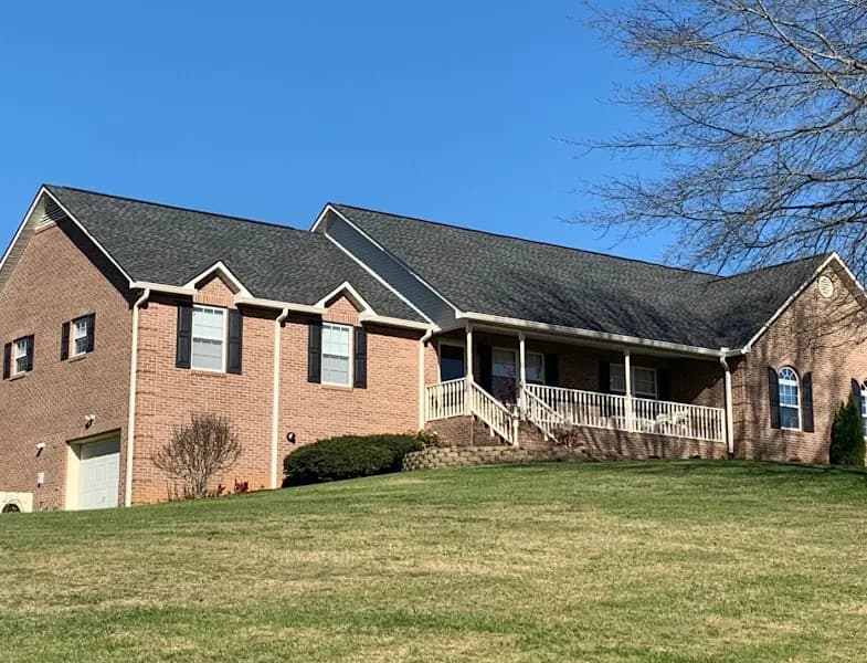 A brick single-story home with a dark asphalt shingle roof, front porch, and well-maintained lawn under a clear blue sky