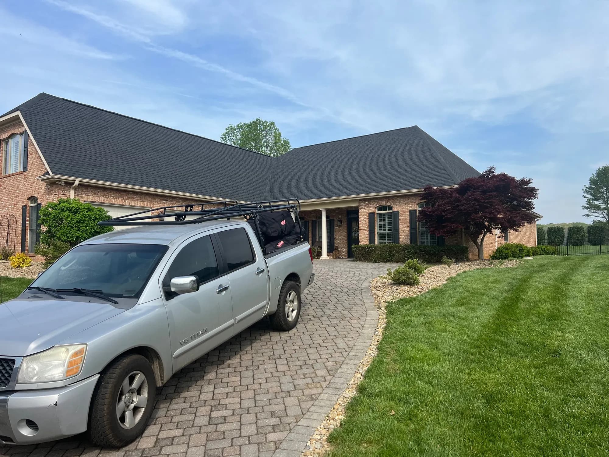 A pickup truck with a ladder rack parked in the driveway of a large brick home with a newly installed dark shingle roof