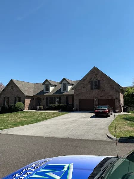 A large brick two-story house with a steep gray shingle roof, viewed from the driveway with a car parked in front of the garage