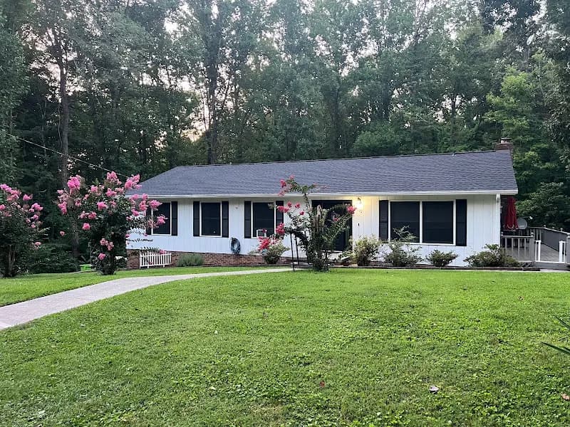 A single-story white ranch-style home with a gray shingle roof, surrounded by lush green lawn, flowering shrubs, and trees at dusk