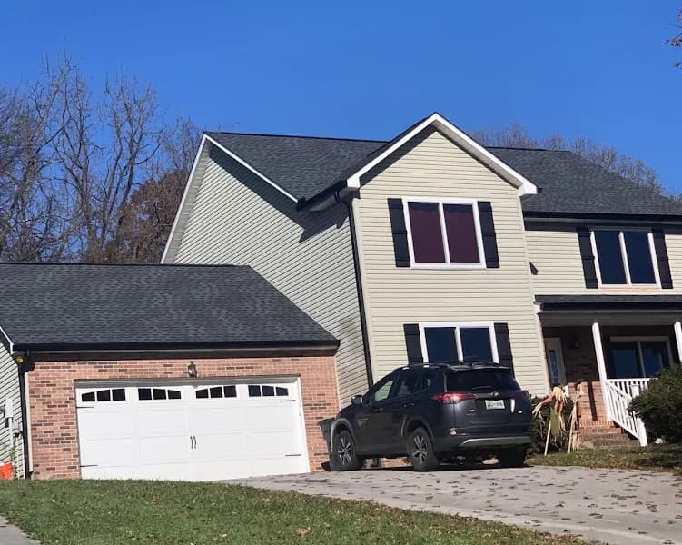 A two-story house with tan siding, brick accents, dark shingle roof, and a two-car garage, with an SUV parked in the driveway