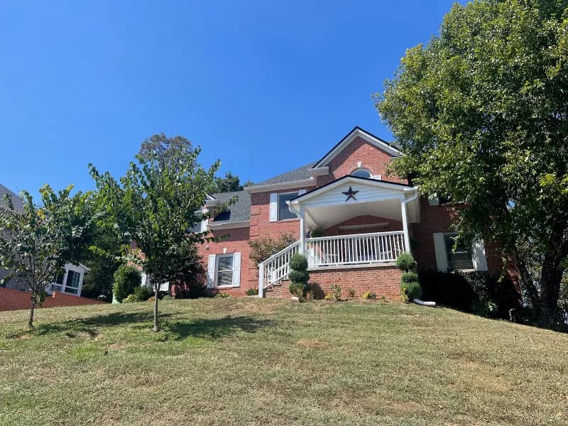 A large brick two-story home with a covered porch featuring a decorative star, surrounded by trees and a green lawn under a clear blue sky
