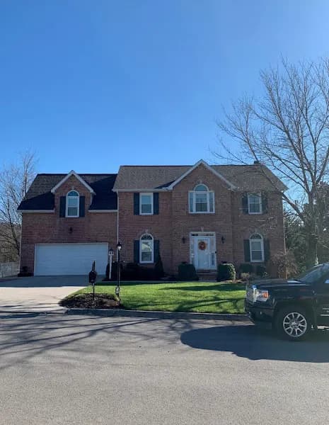 A two-story brick colonial-style home with a black shingle roof, dark shutters, and a two-car garage, photographed from the street on a clear day