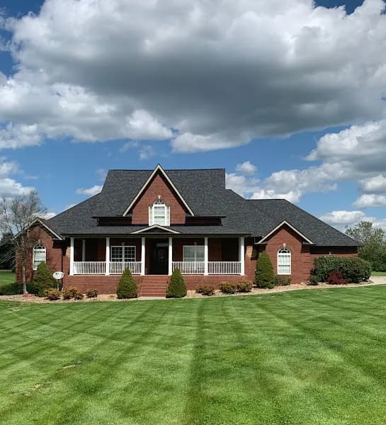 A brick two-story home with a dark shingled roof, covered front porch, and manicured green lawn under a partly cloudy sky