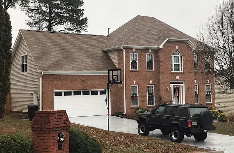 A two-story brick and vinyl-sided home with a gray shingle roof, holiday wreaths on the windows, and a black SUV parked in the driveway