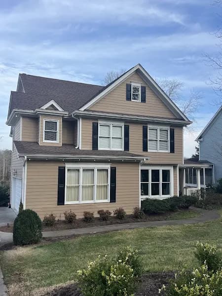 A two-story tan house with dark shutters, a well-maintained roof, and a two-car garage, photographed from the street on a partly cloudy day