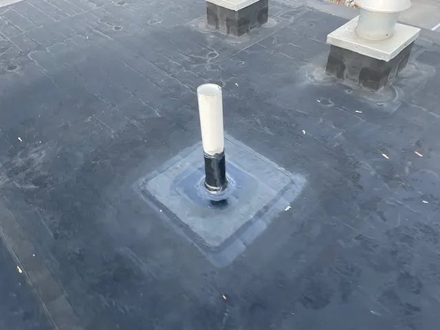Pipe penetration sealed with a square membrane patch on a black EPDM commercial roof, rooftop vents behind