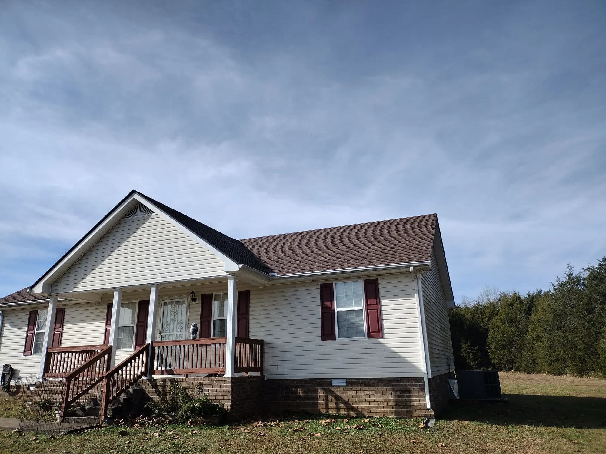 A single-story house with beige vinyl siding, a newly installed brown shingle roof, and a front porch with maroon shutters and steps