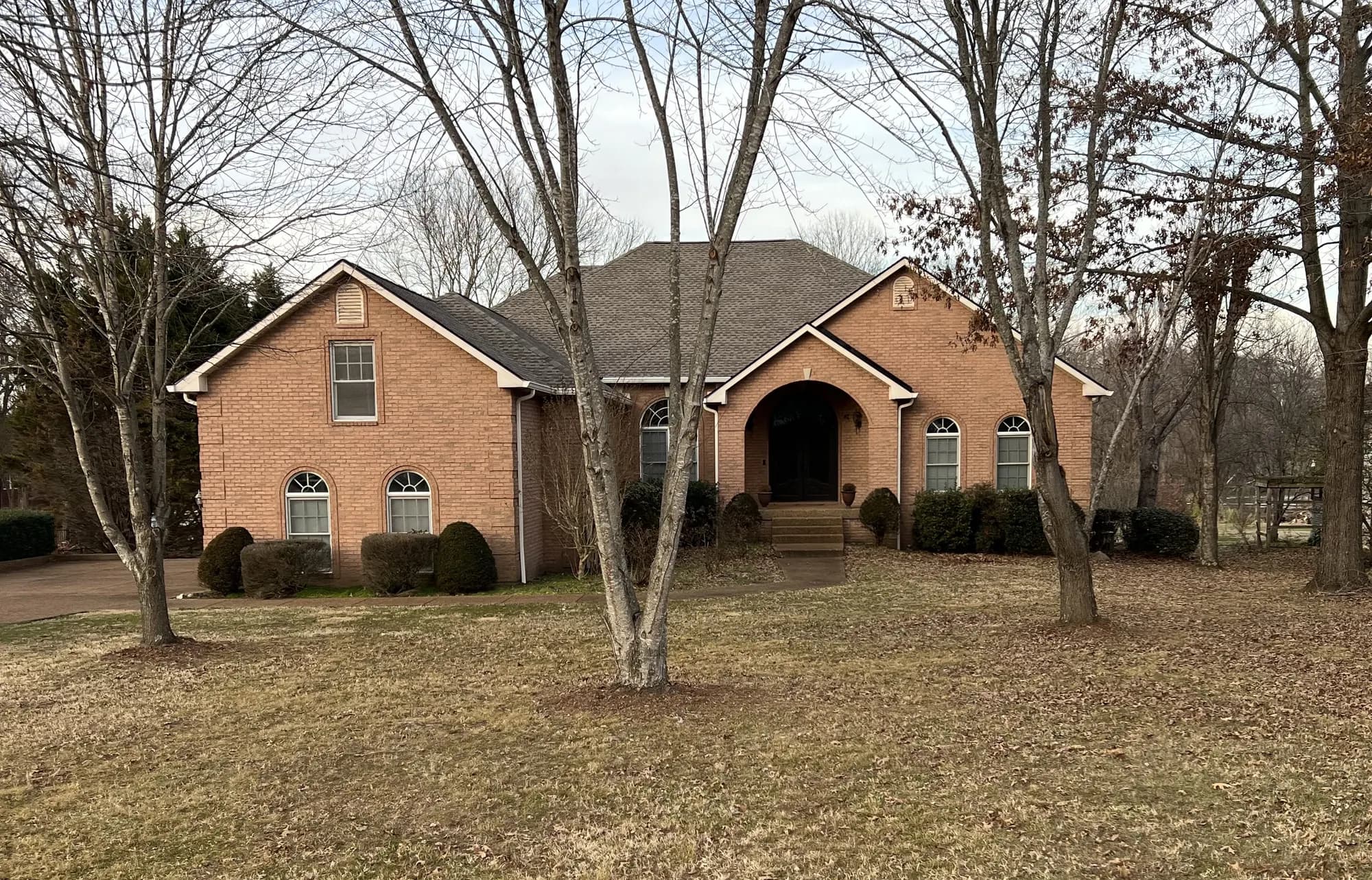 A two-story brick house with an arched entryway and gray shingle roof, surrounded by bare trees in a residential yard