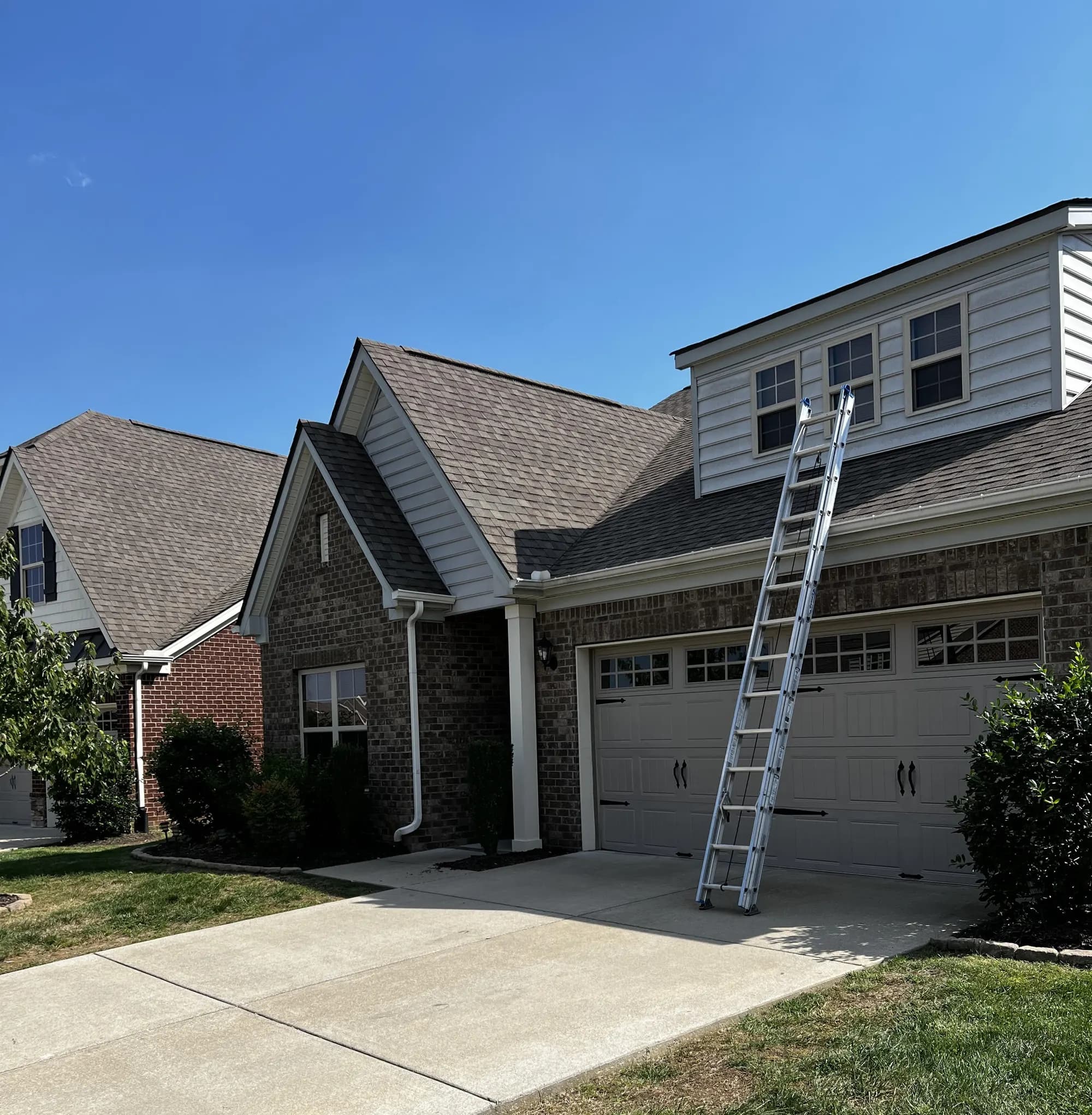 A two-story brick and vinyl-sided home with an extension ladder set up against the roofline near a dormer window, under a clear blue sky