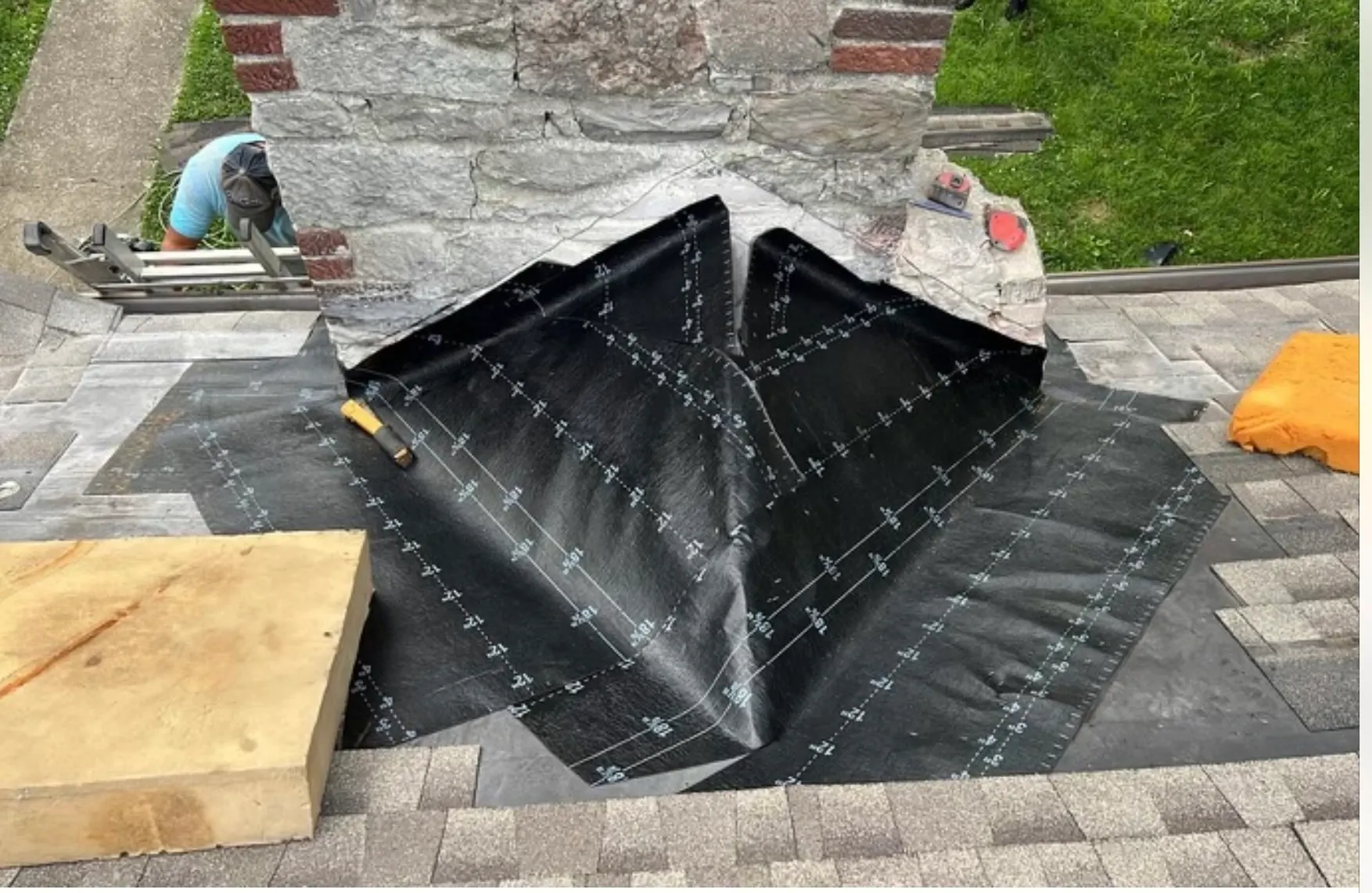 Roofing worker installing waterproof underlayment around a stone chimney during a roof valley repair, viewed from above