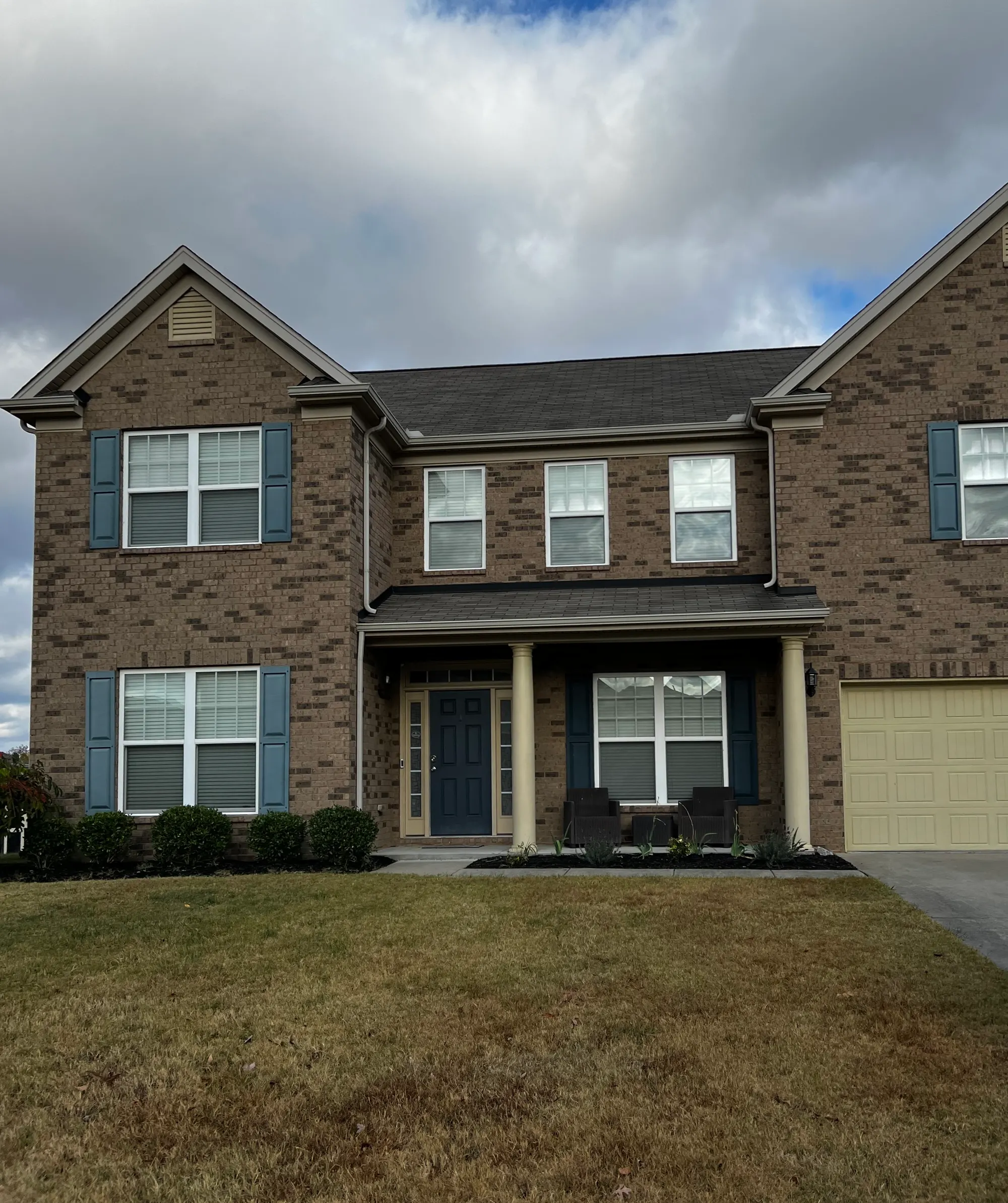 A two-story brick house with blue shutters, a covered front porch, and a manicured lawn under a cloudy sky