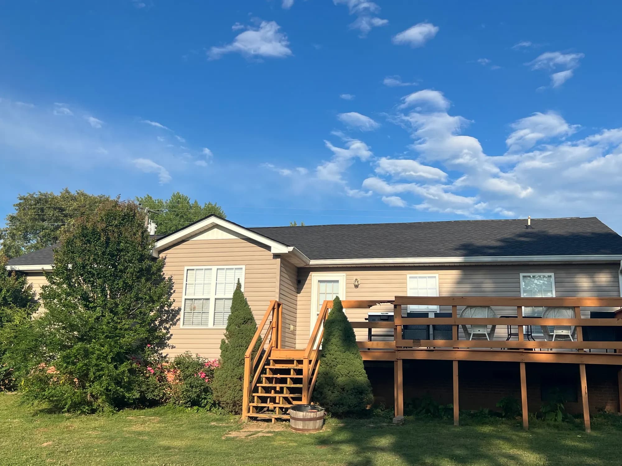 A single-story home with tan vinyl siding, a dark shingle roof, and a wooden deck with outdoor seating, set against a blue sky