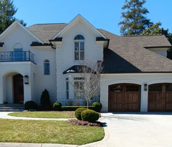 A large upscale two-story stucco home with a slate-style shingle roof, arched entryway, balcony, and attached three-car garage with wood-panel doors