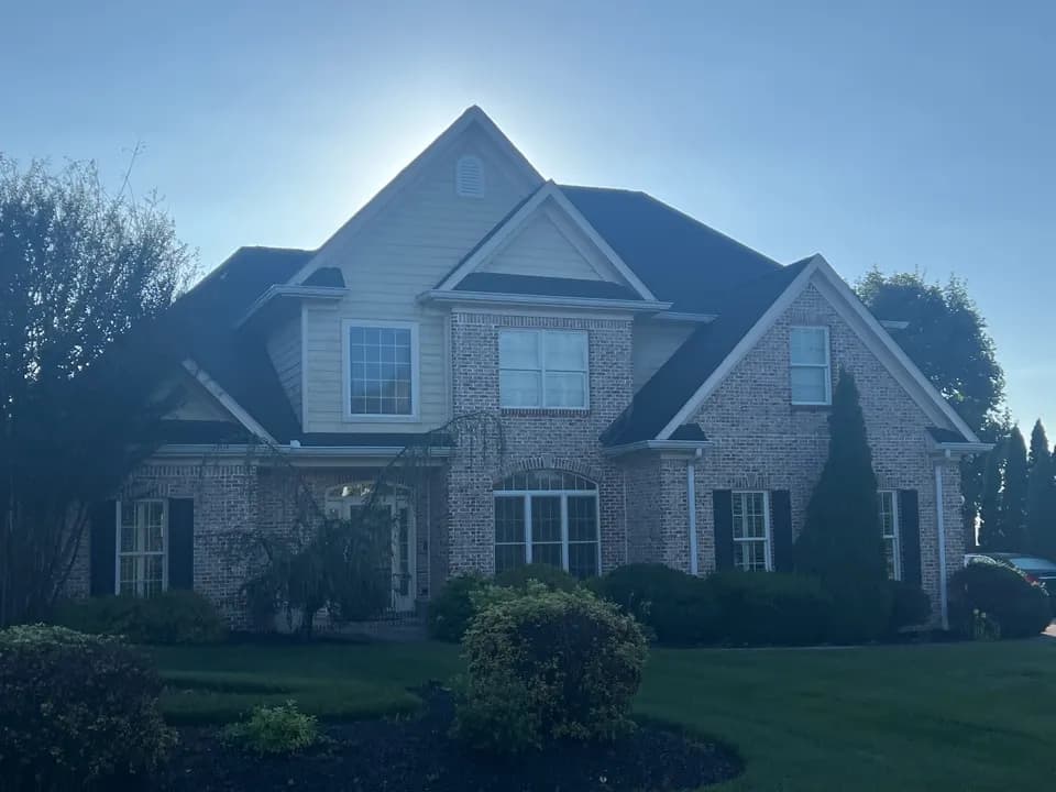 A two-story brick and vinyl-sided home with a dark asphalt shingle roof, photographed from the front lawn with the sun peeking over the roofline