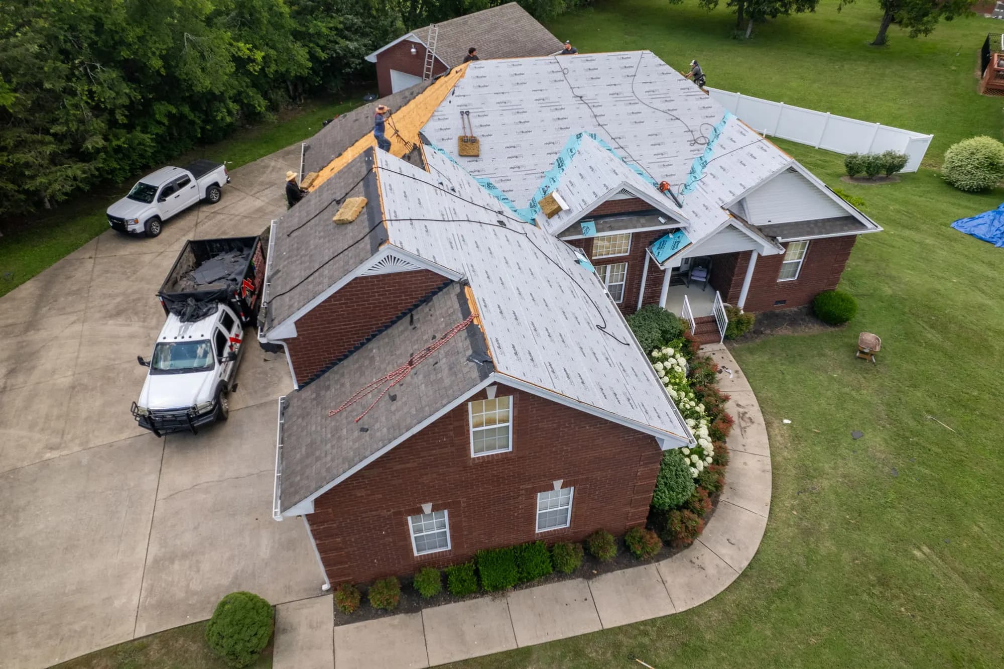 Aerial drone view of roofers installing underlayment on a large brick house during a roof replacement, with a work truck and debris trailer in the driveway