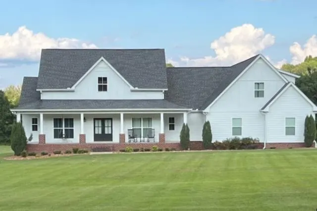 A two-story white farmhouse-style home with a gray shingle roof, covered front porch, and brick accents, surrounded by a manicured lawn