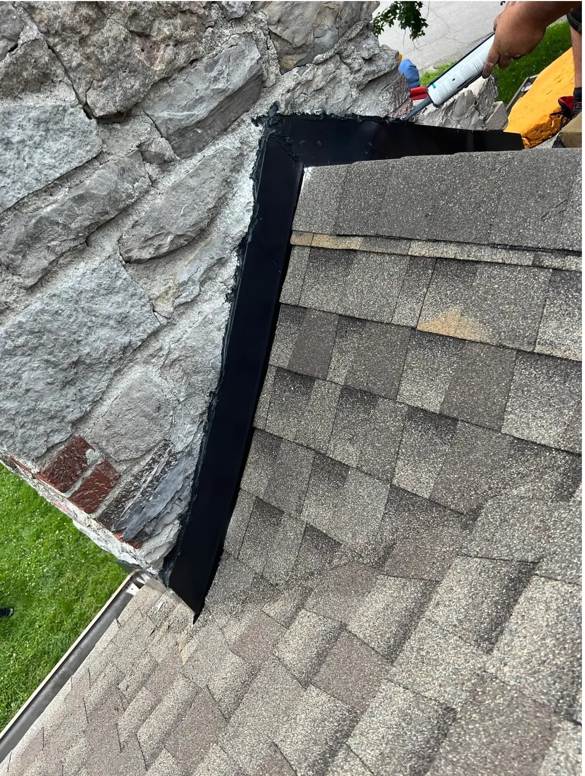 Close-up of newly installed roof flashing where a shingle roof meets a stone chimney, with a worker applying sealant