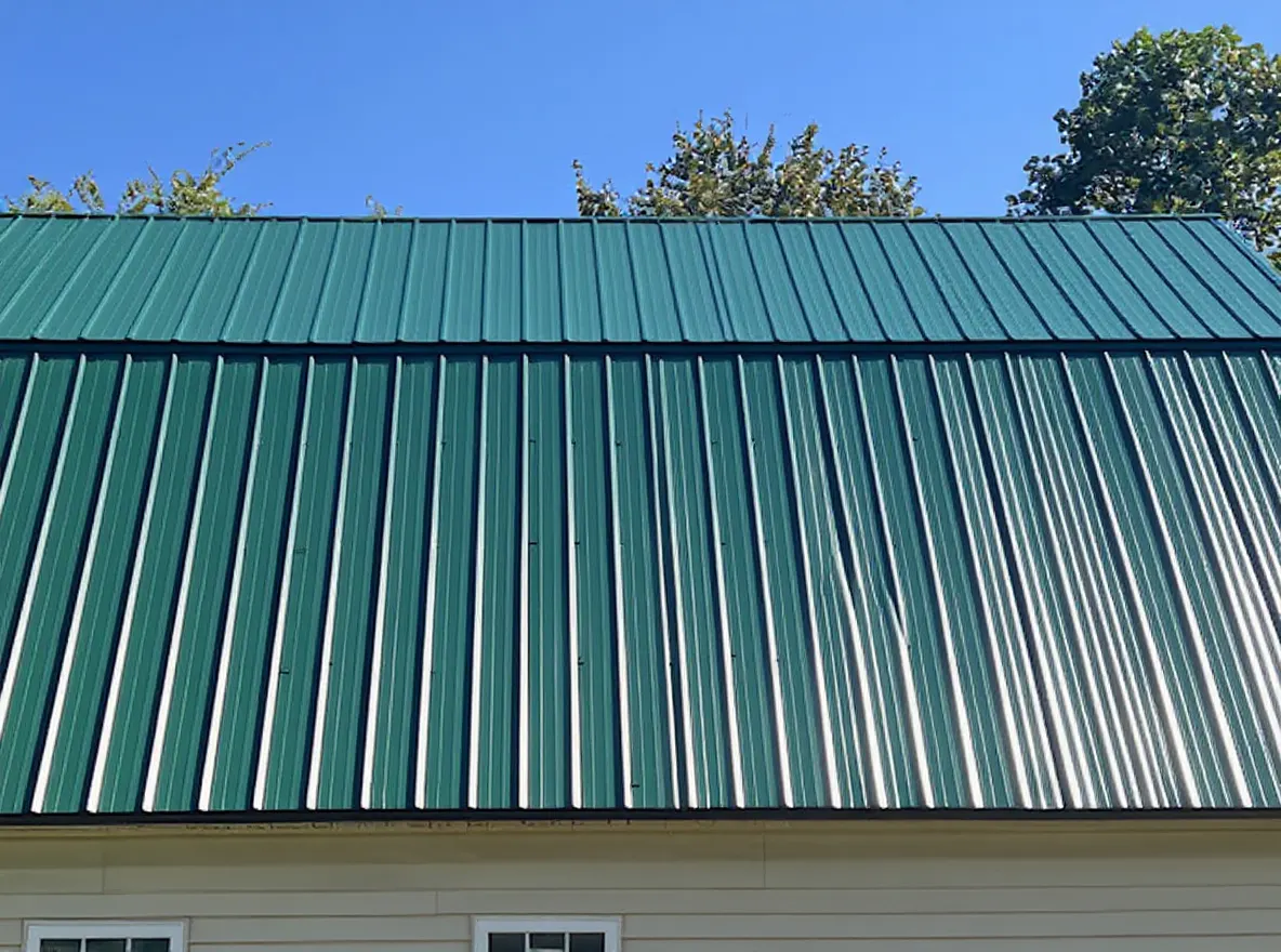 A building with beige siding and a newly installed green metal standing-seam roof, with a red tractor and lawn equipment parked in front