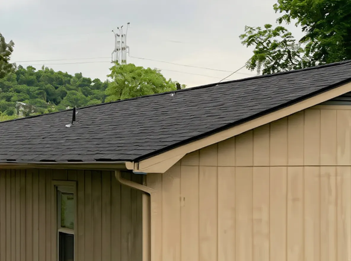 Exterior view of a tan single-story house with a dark shingle roof, wooden deck, and chain-link fence, surrounded by trees and lawn