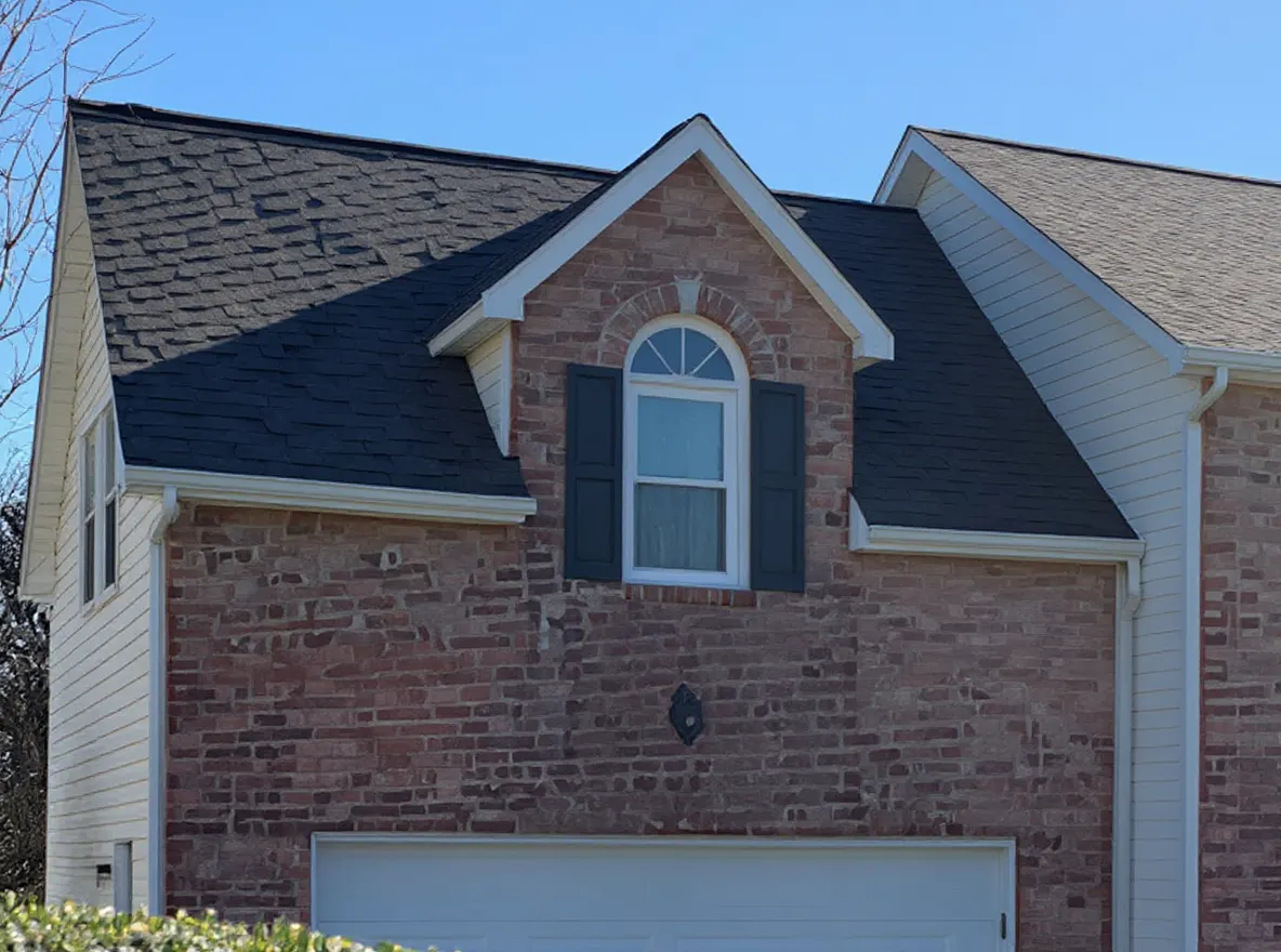 Closer view of the roof: a two-story brick and vinyl-siding home with a gray shingle roof, attached two-car garage, and landscaped front yard on a sunny day