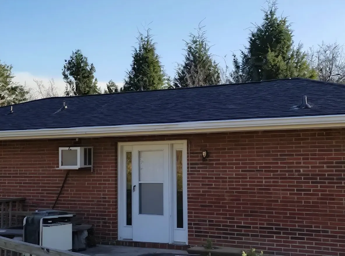 Rear exterior view of a one-story brick ranch home showing a newly installed dark shingle roof, wooden deck, and landscaped yard