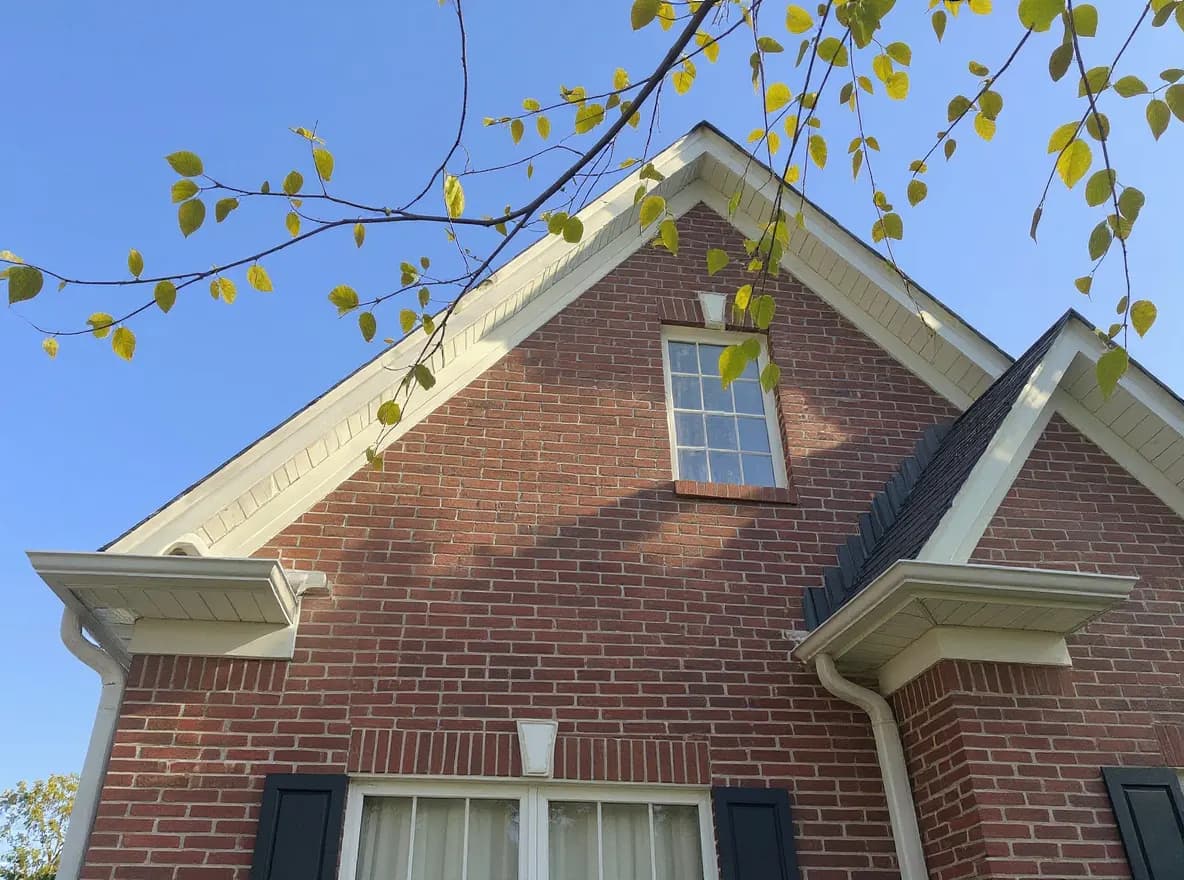 Closer view of the roof: exterior view of a red brick two-story house with a gabled roof, dormer window, and landscaped hedges, framed by tree branches